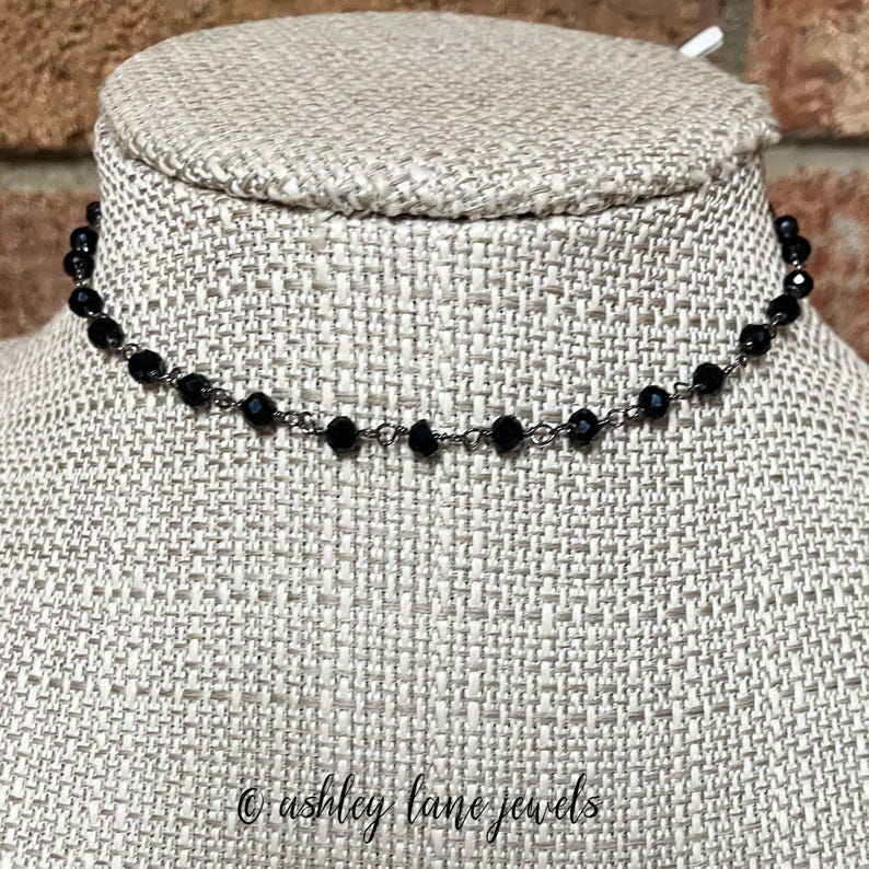May include: A delicate choker necklace featuring small, faceted black beads strung along a thin, dark metallic chain. The necklace is displayed on a textured, light-colored fabric form. The text ashley lane jewels is visible at the bottom.