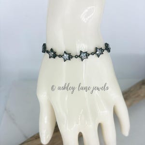 May include: A dark metal chain bracelet featuring a series of connected star-shaped charms, each set with a clear, sparkling stone in the center. The text ashley lane jewels is visible below the bracelet on the display form.