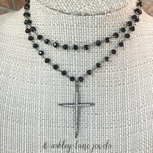 May include: A two-strand necklace with black beads and a cross pendant. The cross is encrusted with small, clear crystals. The necklace is displayed on a neutral-colored surface. The text "ashley lane jewels" is at the bottom.