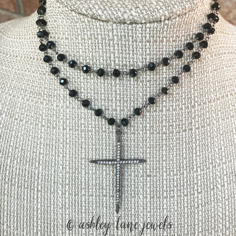 May include: A two-strand necklace with black beads and a cross pendant. The cross is encrusted with small, clear crystals. The necklace is displayed on a neutral-colored surface. The text "ashley lane jewels" is at the bottom.