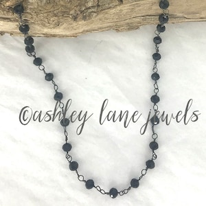 May include: A necklace featuring small, faceted black beads linked together with dark metal wire. The beads are spaced evenly along the chain, which is draped in a U-shape. The text "ashley lane jewels" is printed in black script across the center.