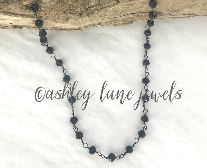 May include: A necklace featuring small, faceted black beads linked together with dark metal wire. The beads are spaced evenly along the chain, which is draped in a U-shape. The text "ashley lane jewels" is printed in black script across the center.