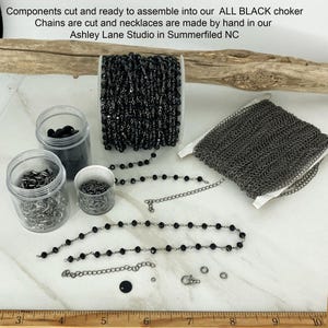 May include: Components for an all black choker including a spool of black beaded chain, a spool of dark metal link chain, three clear plastic jars containing metal clasps and rings, and loose chain segments with a ruler showing 10 inches.