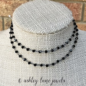 May include: A double-strand necklace featuring small, faceted black beads strung on a dark metal chain. The necklace is displayed on a textured, light-colored form. The text "ashley lane jewels" is printed in a cursive font at the bottom.