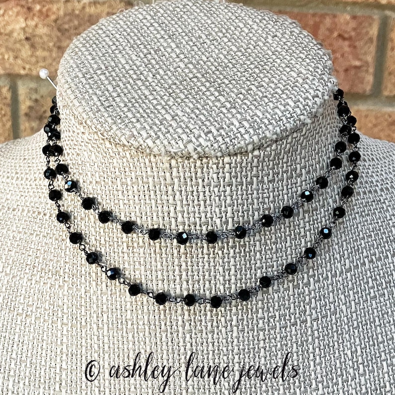 May include: A double-strand necklace featuring small, faceted black beads strung on a dark metal chain. The necklace is displayed on a textured, light-colored form. The text "ashley lane jewels" is printed in a cursive font at the bottom.