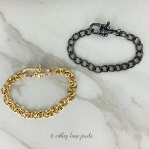 May include: Two chain bracelets, one gold and one black, with a clasp closure. The bracelets are on a white marble surface.