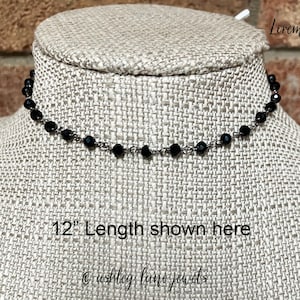 May include: A black beaded choker necklace featuring small, faceted, round beads spaced evenly along a thin metal chain. The necklace is displayed on a textured form with the text 12" Length shown here and ashley lane jewels printed on it.