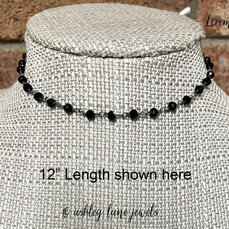 May include: A black beaded choker necklace featuring small, faceted, round beads spaced evenly along a thin metal chain. The necklace is displayed on a textured form with the text 12" Length shown here and ashley lane jewels printed on it.