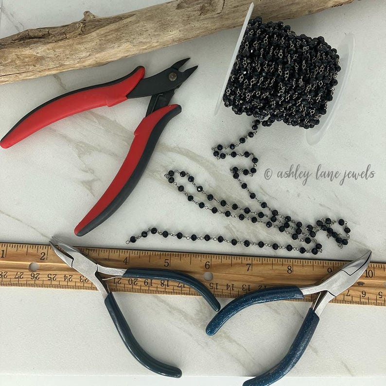May include: A spool of black beaded chain, a pair of red and black wire cutters, a pair of blue-handled pliers, and a wooden ruler measuring 10 inches. The text "ashley lane jewels" is visible on the surface near the beaded chain.