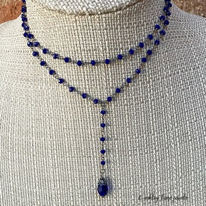 May include: A two-strand necklace with a Y-shaped pendant. The necklace features small, round, dark blue beads strung on a silver-colored chain. The pendant has a teardrop-shaped blue gemstone at the end. Jewelry.