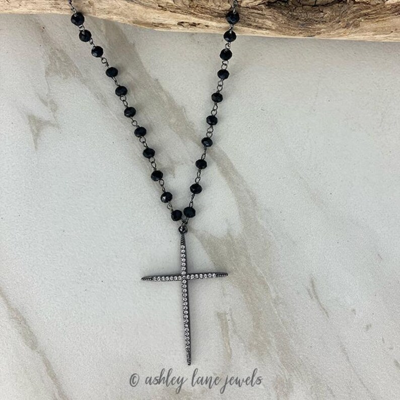 May include: A black beaded necklace with a silver cross pendant. The cross is encrusted with small, clear crystals. The necklace is displayed on a light-colored surface with a wooden accent.