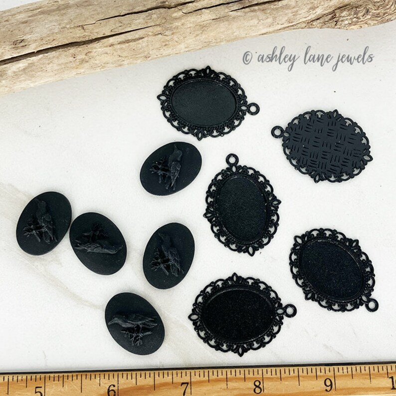 May include: A collection of eight black metal cameo pendants, including six with intricate filigree designs and two with raised bird motifs.  The pendants are arranged on a white surface with a wooden ruler in the foreground.