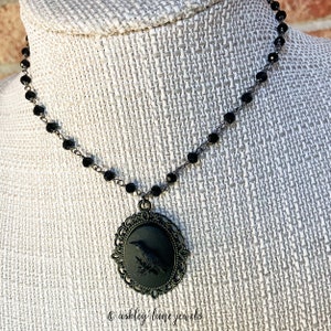 May include: A black choker necklace with a black cameo pendant featuring a raven silhouette. The cameo is set in an ornate, filigree frame.