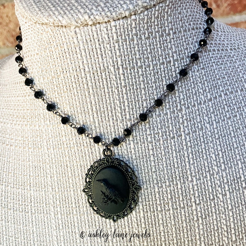 May include: A black choker necklace with a black cameo pendant featuring a raven silhouette. The cameo is set in an ornate, filigree frame.