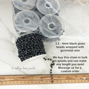 May include: A spool of black glass beads wrapped with gunmetal wire, measuring 3.5 - 4mm. The text reads: 3.5 - 4mm black glass beads wrapped with gunmetal wire. We buy this chain in bulk on spools and can make any length you need. Message us for a custom order.