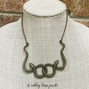 May include: A gold-toned necklace featuring two intertwined snake figures. The snakes are facing each other and their tails are joined together. The necklace is on a mannequin's neck.