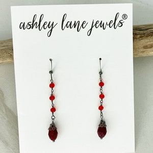 May include: A pair of dangle earrings with a red teardrop-shaped crystal at the bottom. The earrings are made with a silver chain and red beads.