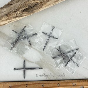 May include: Several silver-toned cross-shaped jewelry pendants, each encrusted with small, sparkling clear stones. The pendants are packaged in clear plastic bags. A wooden ruler is visible at the bottom of the image.