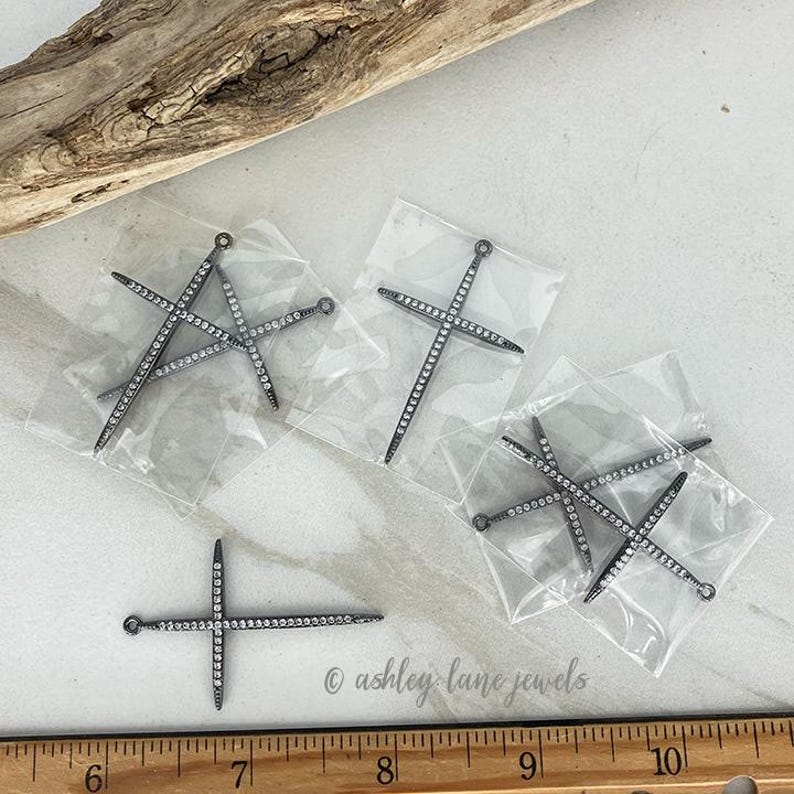 May include: Several silver-toned cross-shaped jewelry pendants, each encrusted with small, sparkling clear stones. The pendants are packaged in clear plastic bags. A wooden ruler is visible at the bottom of the image.