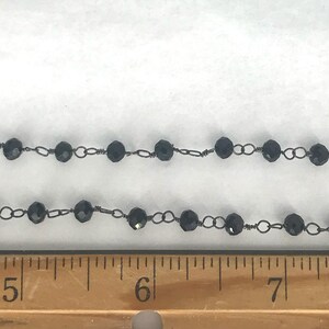 May include: A black beaded chain featuring faceted round beads linked by thin dark metal wire, positioned above a wooden ruler showing measurements in inches, with the beads spanning from the 4.5-inch mark to beyond the 7.5-inch mark.