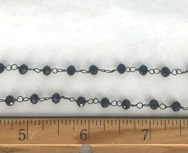May include: A black beaded chain featuring faceted round beads linked by thin dark metal wire, positioned above a wooden ruler showing measurements in inches, with the beads spanning from the 4.5-inch mark to beyond the 7.5-inch mark.