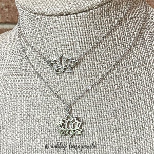 May include: A silver chain necklace with two delicate lotus flower pendants. The necklace is displayed on a textured fabric background.