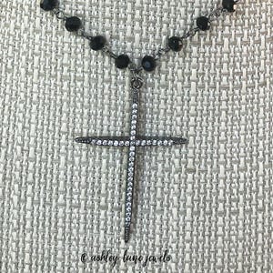 May include: A black beaded necklace with a pendant featuring a cross shape. The cross is encrusted with small, sparkling white stones. The necklace is displayed against a textured, light-colored background. The text "@ashley lane jewels" is at the bottom.