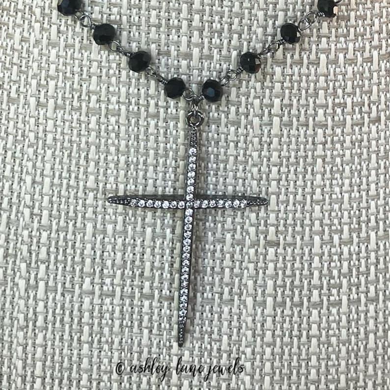 May include: A black beaded necklace with a pendant featuring a cross shape. The cross is encrusted with small, sparkling white stones. The necklace is displayed against a textured, light-colored background. The text "@ashley lane jewels" is at the bottom.