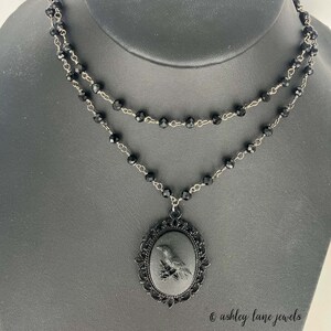 May include: A black necklace with a black cameo pendant featuring a raven. The necklace is made of black beads and silver chain.