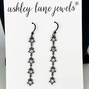May include: A pair of black dangle earrings with a chain of silver star-shaped charms. The earrings are on a white card with the text "ashley lane jewels" in black cursive.