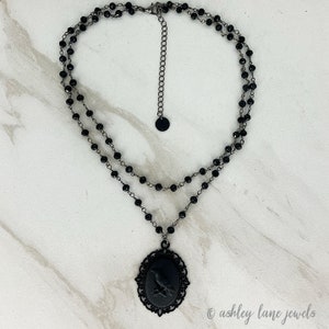 May include: A black choker necklace with a black cameo pendant featuring a raven. The necklace is made of two strands of black beads.