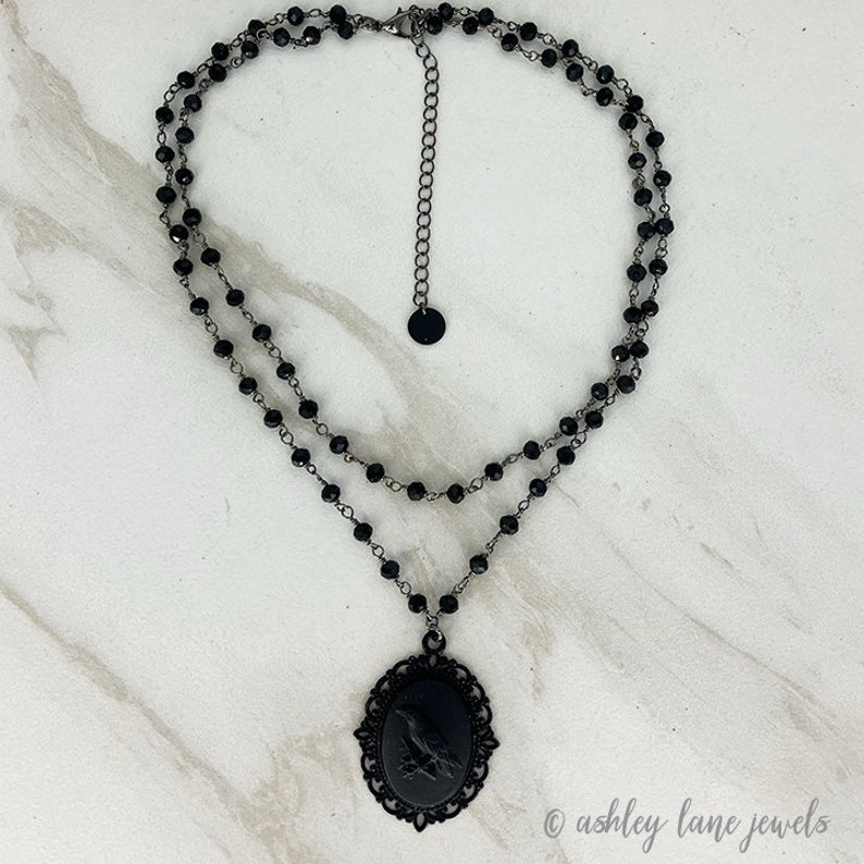 May include: A black choker necklace with a black cameo pendant featuring a raven. The necklace is made of two strands of black beads.