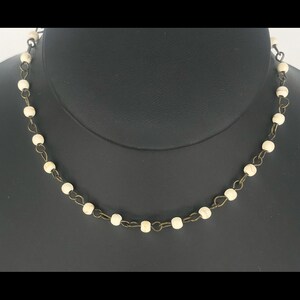 May include: A delicate necklace featuring small, off-white beads strung together with a dark, antique-style chain. The beads are evenly spaced, creating a simple yet elegant design. The necklace is displayed against a dark background, highlighting its details.