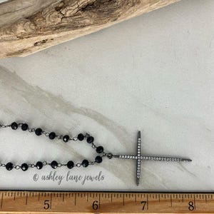 May include: A black beaded necklace with a silver cross pendant. The cross is encrusted with small, clear stones. The necklace is displayed on a light-colored surface with a wooden accent.