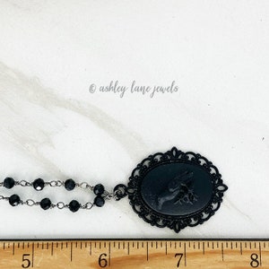 May include: Black necklace with a black cameo pendant featuring a bird design. The pendant is set in a black filigree frame. The necklace is on a white surface with a ruler.