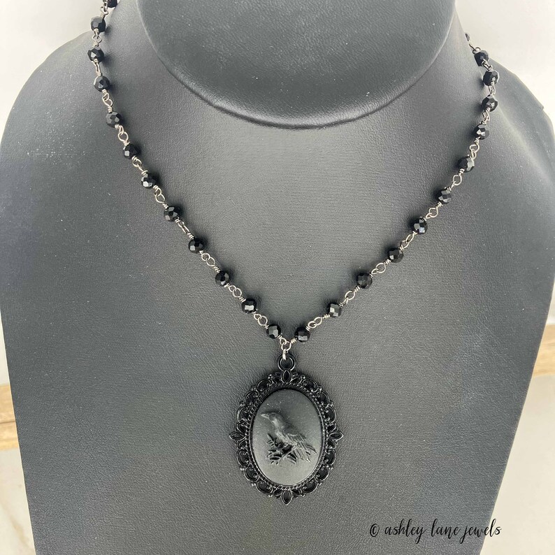 May include: A black choker necklace with a black cameo pendant featuring a raven. The cameo is set in a black metal frame with an ornate design.