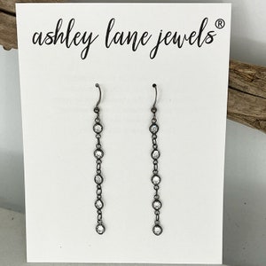 May include: A pair of long, silver dangle earrings with clear crystal stones. The earrings are on a white card with the text "ashley lane jewels" printed on it.
