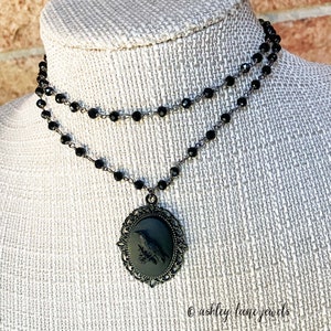May include: A black choker necklace with a black cameo pendant featuring a raven. The necklace is made of black beads and a silver chain.