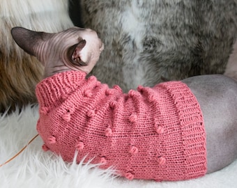 May include: A pink knitted sweater with bobble details, designed for a hairless cat. The sweater is a soft, warm garment. The cat is lying on a white fluffy surface, with a logo in the bottom right corner.