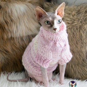 May include: A hairless Sphynx cat wearing a pink cable-knit sweater. The cat is sitting on a white fluffy surface, with a brown and tan faux fur background. The sweater has a high collar and covers the cat's front legs.