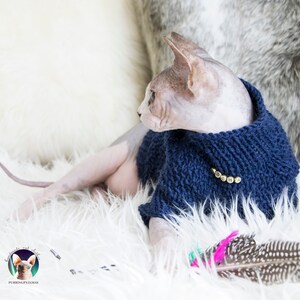 May include: A Sphynx cat wearing a navy blue knitted sweater with gold-colored letters spelling "OSHA". The cat is lying on a white fluffy surface, with a feather toy in the foreground. The image includes a logo for "Purring Pyjamas".