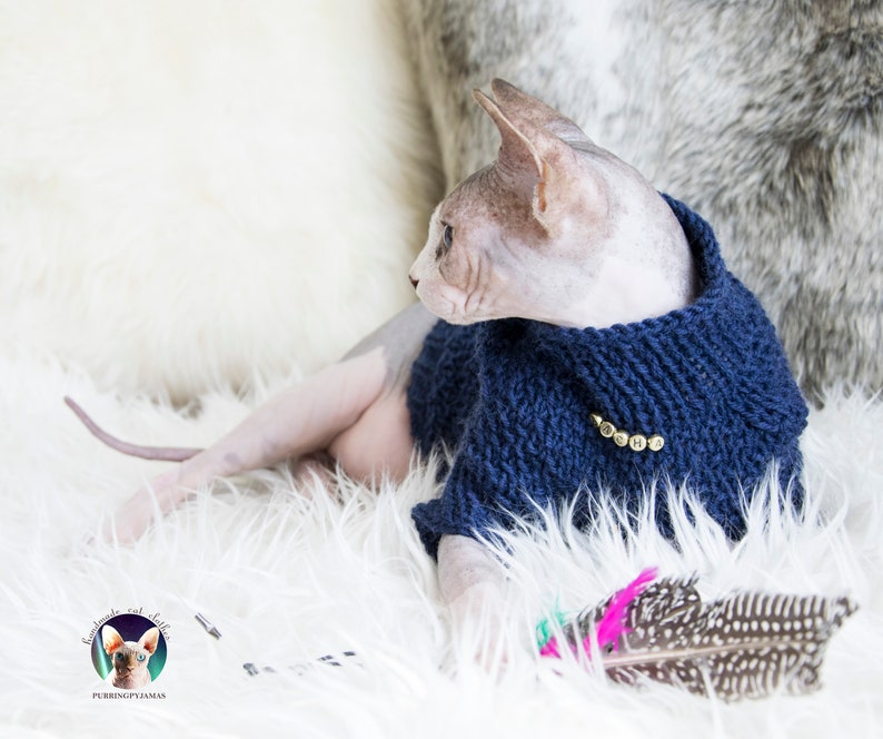 May include: A Sphynx cat wearing a navy blue knitted sweater with gold-colored letters spelling "OSHA". The cat is lying on a white fluffy surface, with a feather toy in the foreground. The image includes a logo for "Purring Pyjamas".