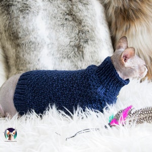May include: A Sphynx cat wearing a navy blue knitted sweater with a high collar. The cat is lying on a white fluffy surface, with a gray and brown furry background. A logo is visible in the bottom left corner.
