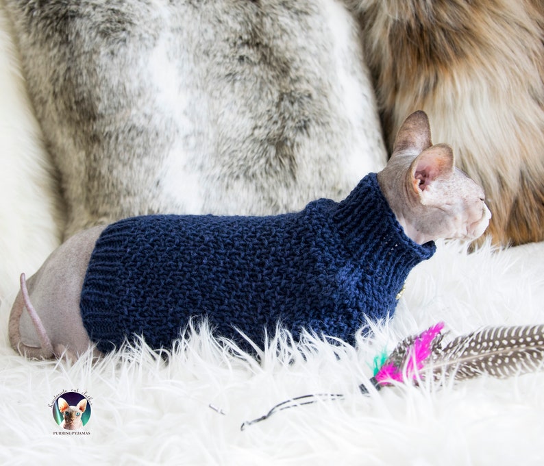 May include: A Sphynx cat wearing a navy blue knitted sweater with a high collar. The cat is lying on a white fluffy surface, with a gray and brown furry background. A logo is visible in the bottom left corner.