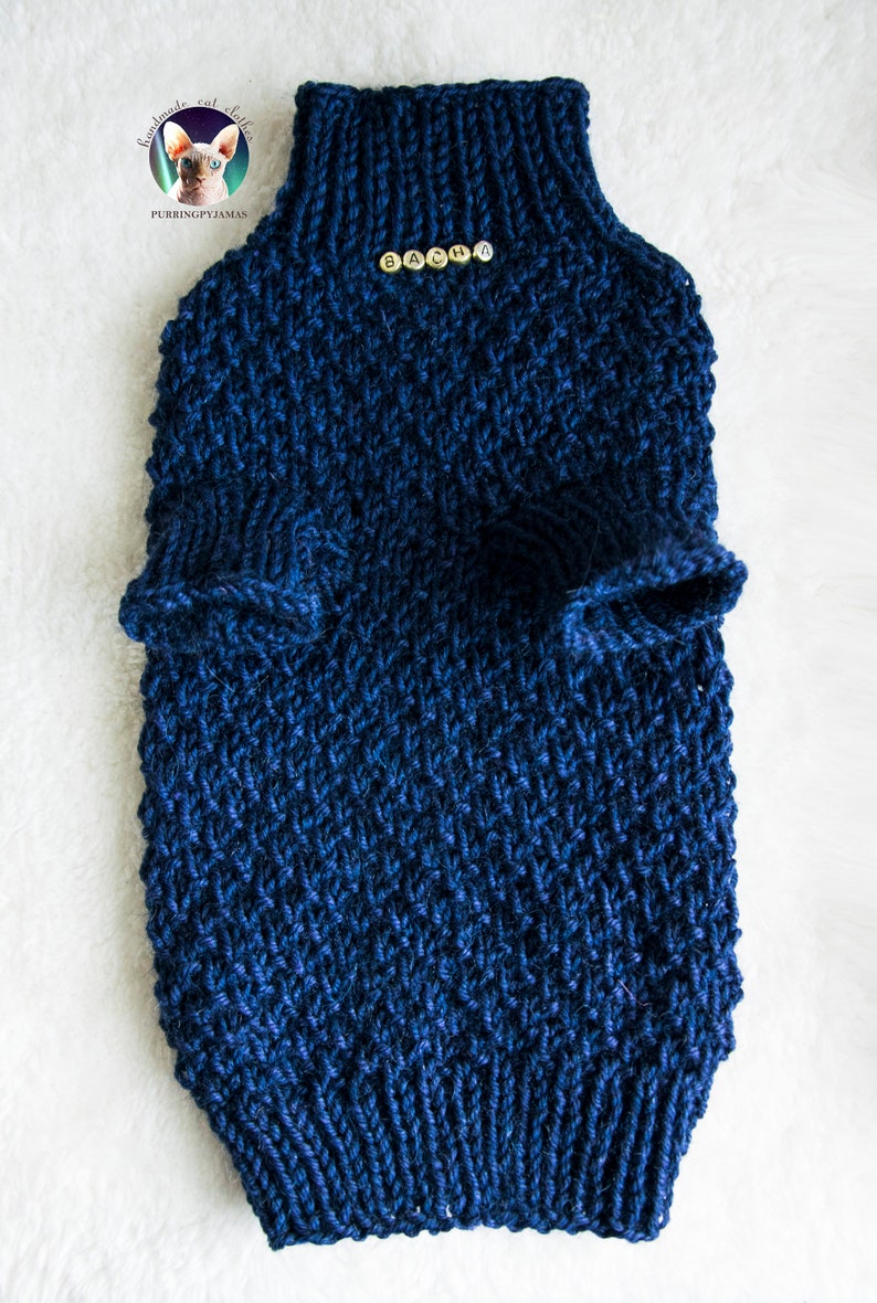May include: A navy blue knitted pet sweater with a high collar and armholes. The sweater has the name "SACHA" spelled out in small gold beads. The item is handmade and designed for cats.