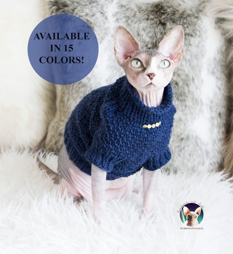 May include: A Sphynx cat wearing a navy blue knitted sweater with a gold-colored embellishment. The sweater has a turtleneck design. The image includes the text "AVAILABLE IN 15 COLORS!" and a logo for "PURRINGPYJAMAS". The cat is sitting on a white fluffy surface.