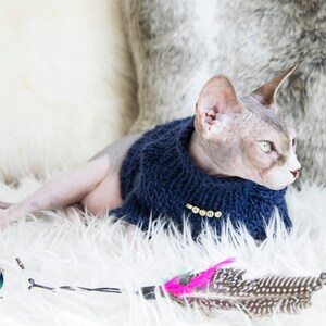 May include: A Sphynx cat wearing a navy blue knitted sweater with gold-colored buttons. The cat is lying on a white fluffy surface with a feather toy nearby. The background includes a white and gray textured surface.