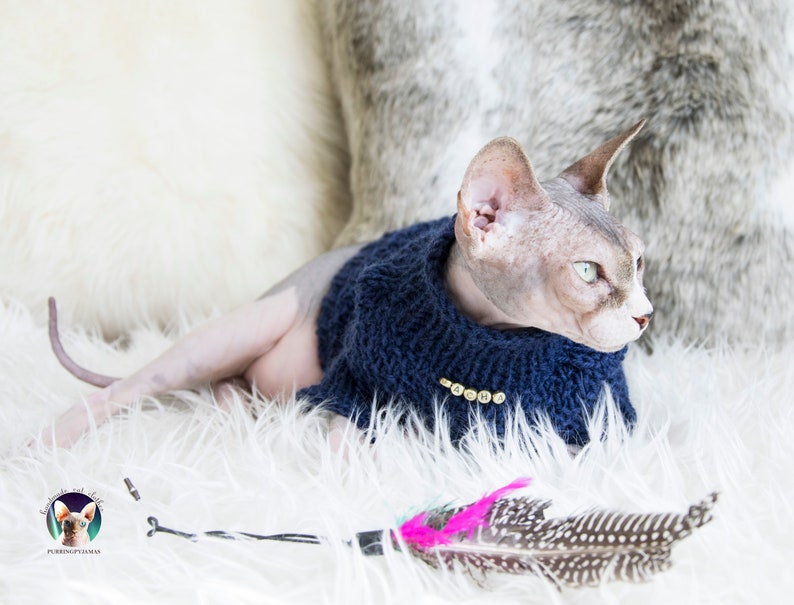May include: A Sphynx cat wearing a navy blue knitted sweater with gold-colored buttons. The cat is lying on a white fluffy surface with a feather toy nearby. The background includes a white and gray textured surface.