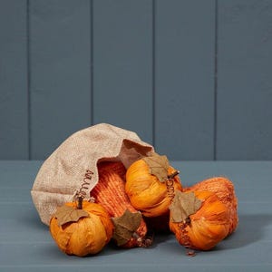 May include: A tan hessian sack spilling out three round orange pumpkins made of textured material. Each pumpkin features a brown paper leaf attached to the top stem. The pumpkins have a ribbed appearance and are arranged in a cluster.