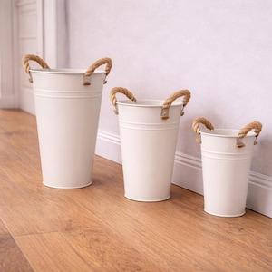 May include: Three white metal buckets of varying sizes with rope handles. The buckets are cylindrical with a slightly flared top and sit on a wooden floor. The rope handles are a natural tan colour.